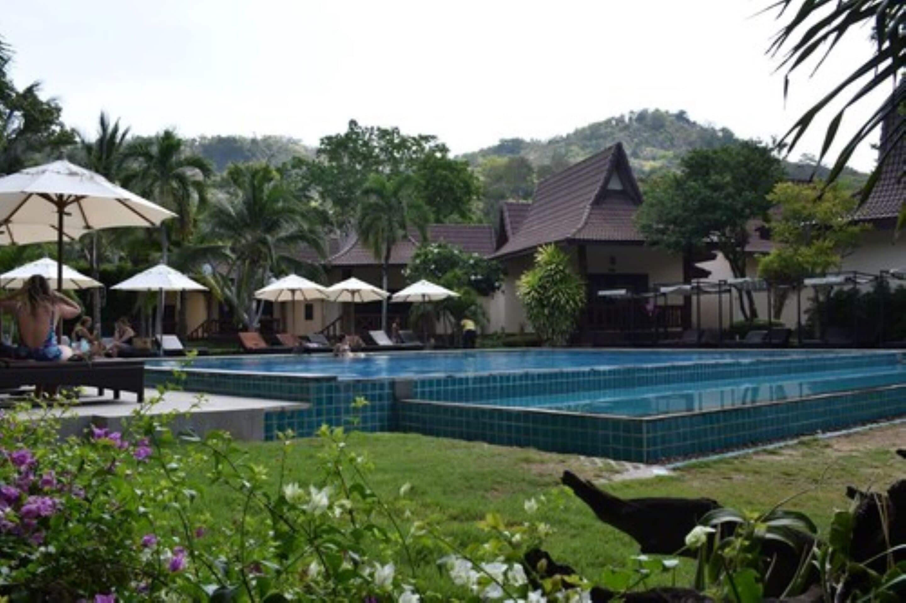 A swimming pool with tiled edges, surrounded by lounge chairs and umbrellas, with a backdrop of tropical vegetation and a hill.