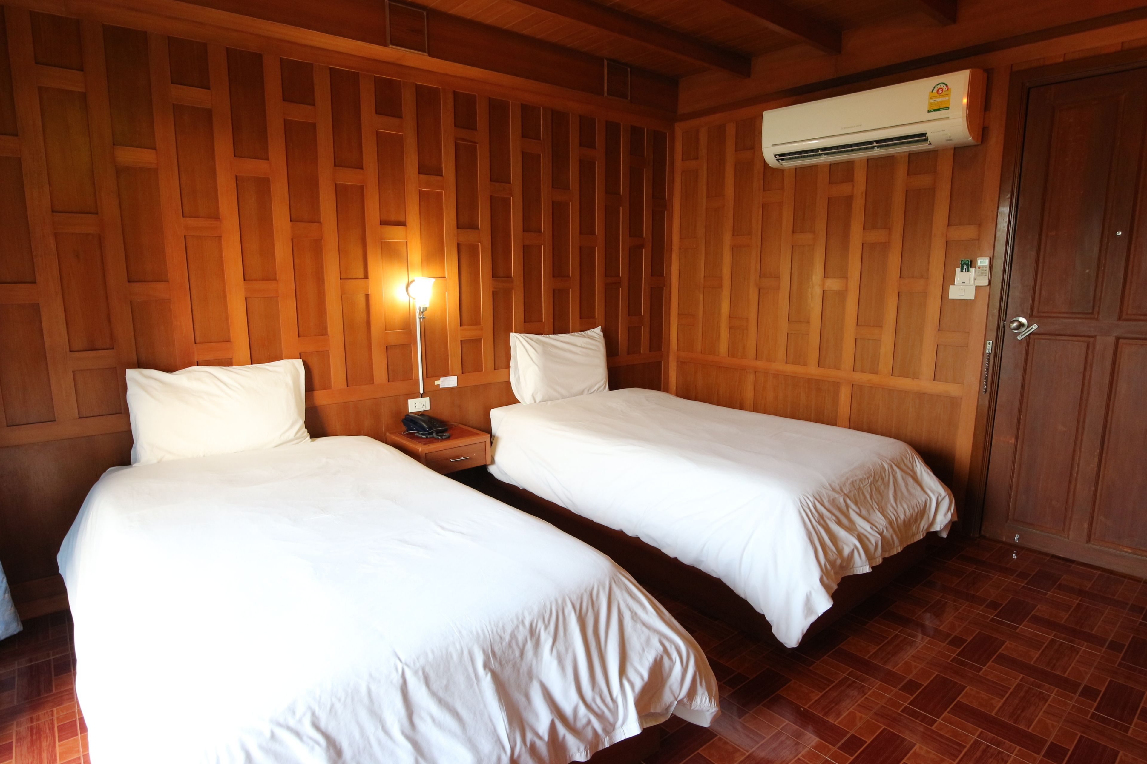 Una habitación con dos camas, paneles de madera en las paredes, un equipo de aire acondicionado, una mesita de noche con una lámpara y una puerta.