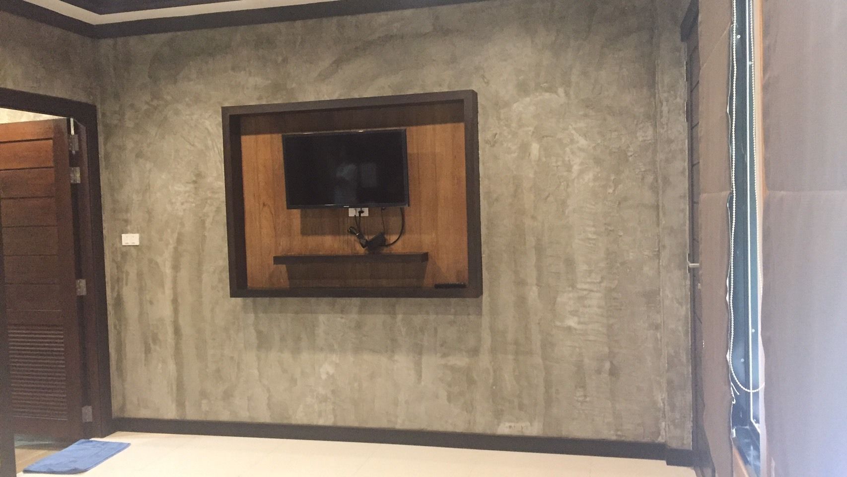 A room with a television mounted on a textured wall, a wooden door, and a window with blinds.