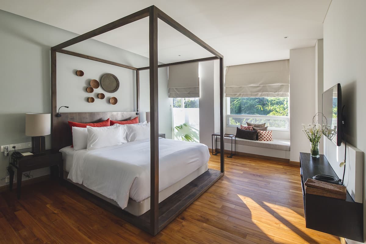 Une chambre moderne avec un lit à baldaquin, une table de chevet avec une lampe, un téléviseur fixé au mur et une fenêtre donnant sur un paysage verdoyant.
