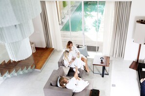 A modern living room with a grey sofa, a wooden coffee table, and a large window with a view of a pool. - Hu'u Villas (Seminyak)