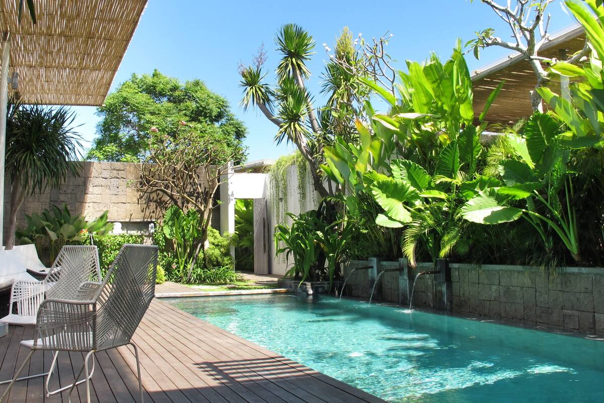 Un espace piscine doté d’une terrasse en bois, de chaises longues et de plantes tropicales.