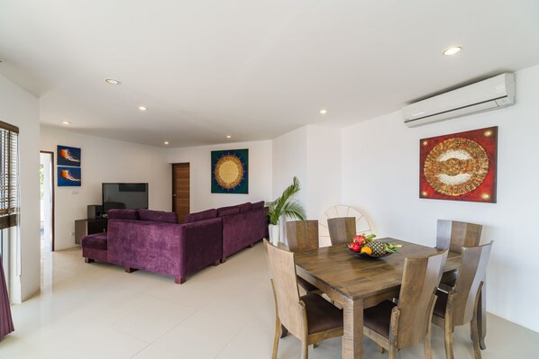 A modern living room with a purple sofa, a wooden dining table with chairs, and a flat-screen TV.
