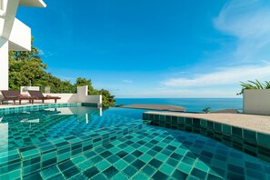 A clear blue infinity pool with a tiled edge, surrounded by lounge chairs and a white building.