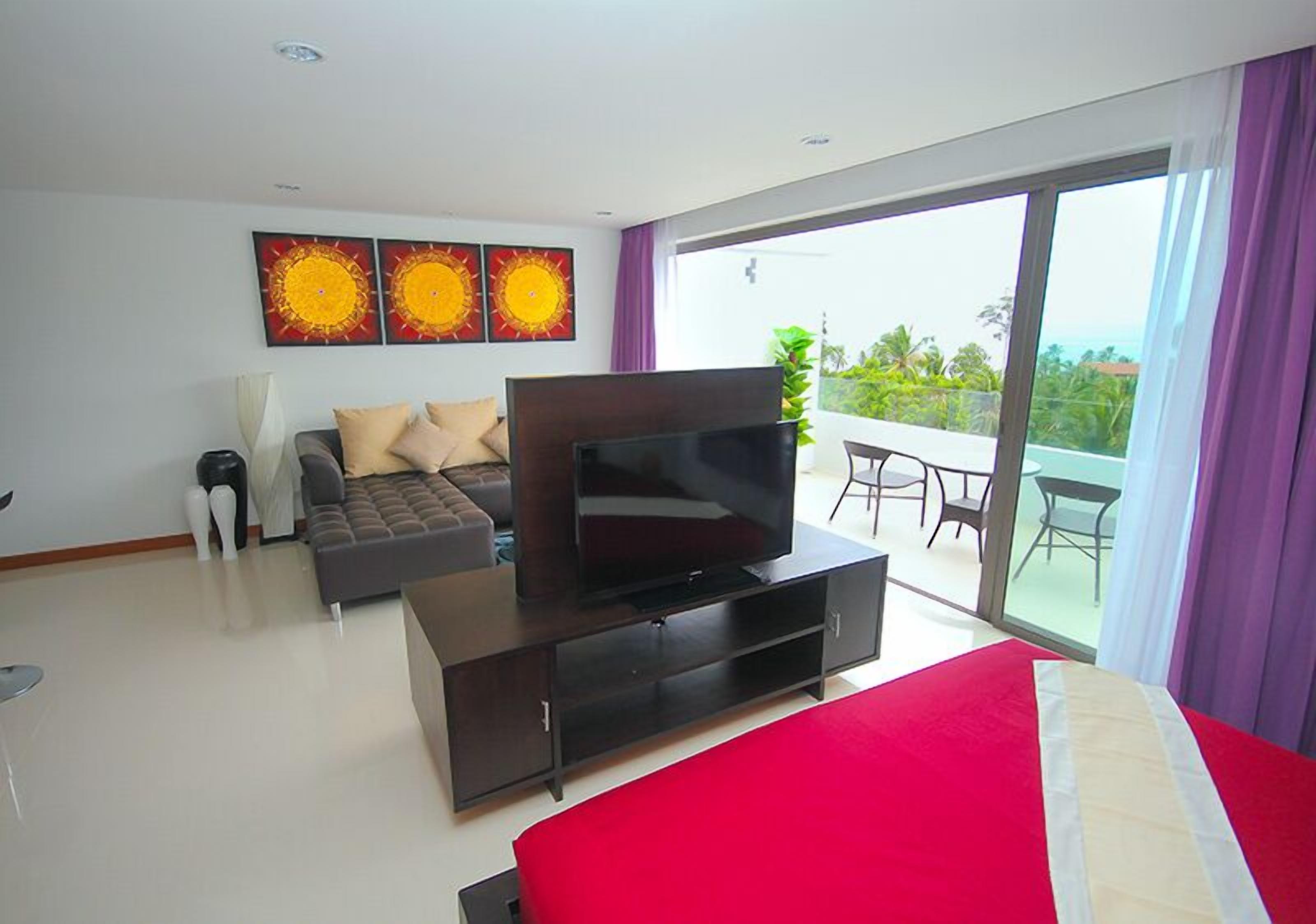 A modern hotel room with a large bed, a TV on a wooden stand, a sofa, and a balcony with a view.