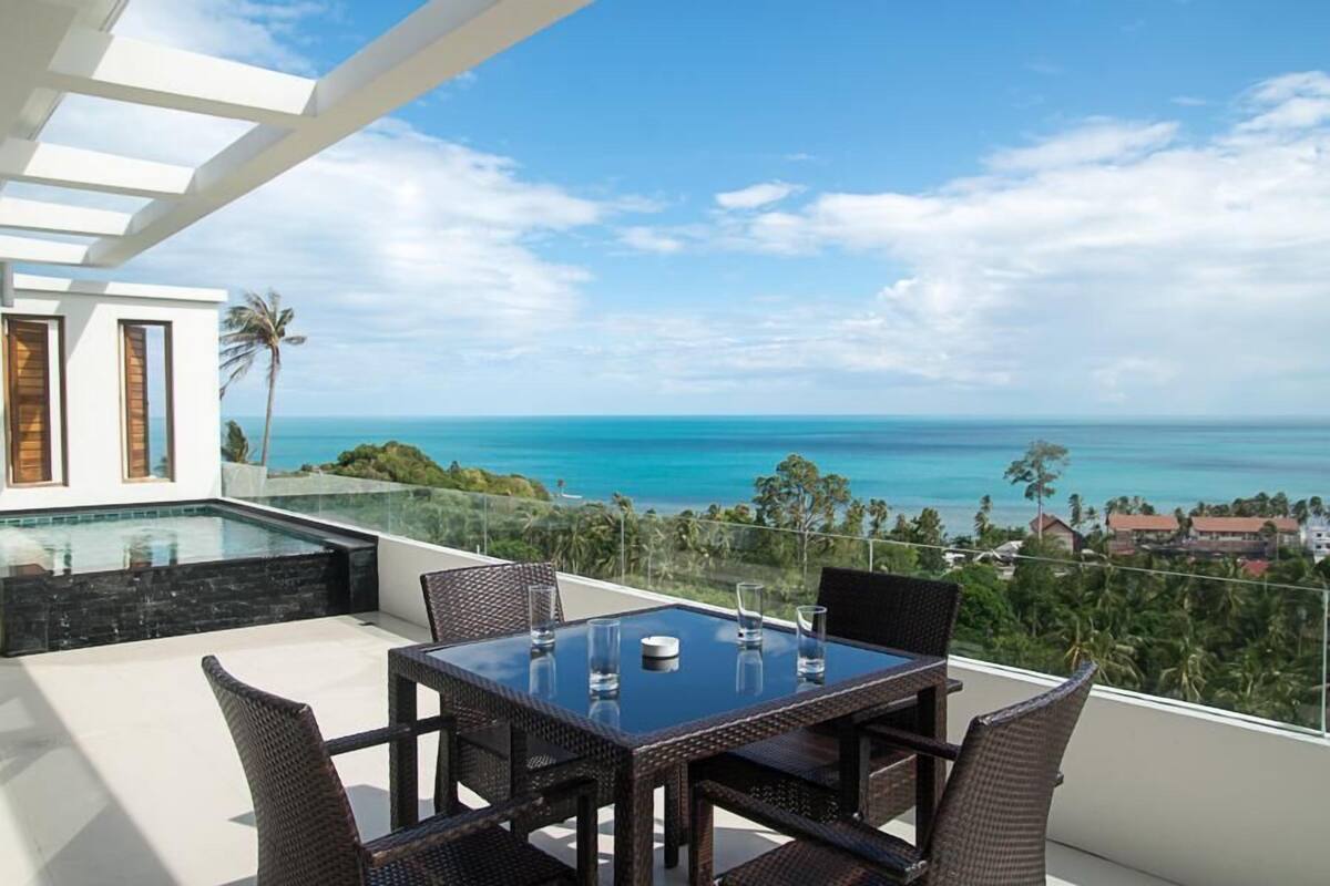 Un balcón con una mesa y sillas de vidrio, con vista al mar, una piscina y palmeras.
