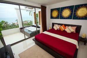 A bedroom with a large bed, a glass-enclosed balcony with a jacuzzi, and a view of the ocean.
