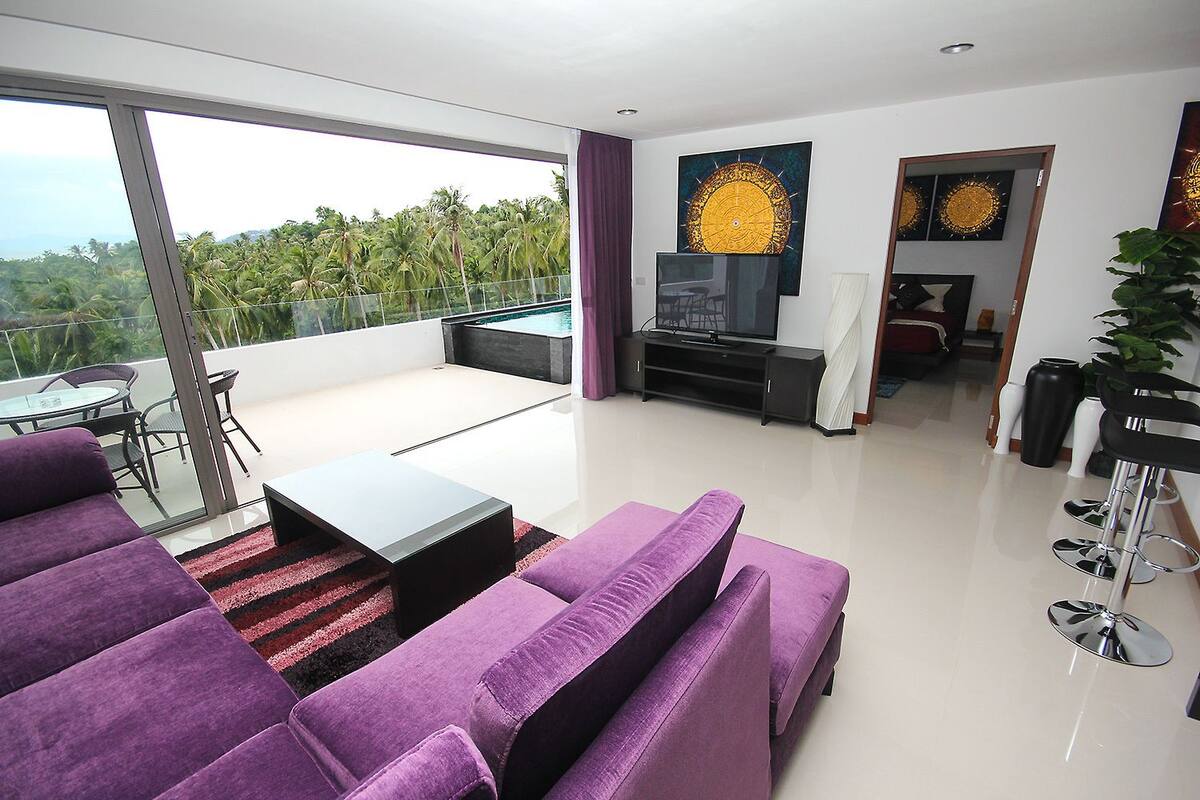 Un salón moderno con un sofá grande, una mesa de centro y un televisor. Hay un balcón con vistas a palmeras.