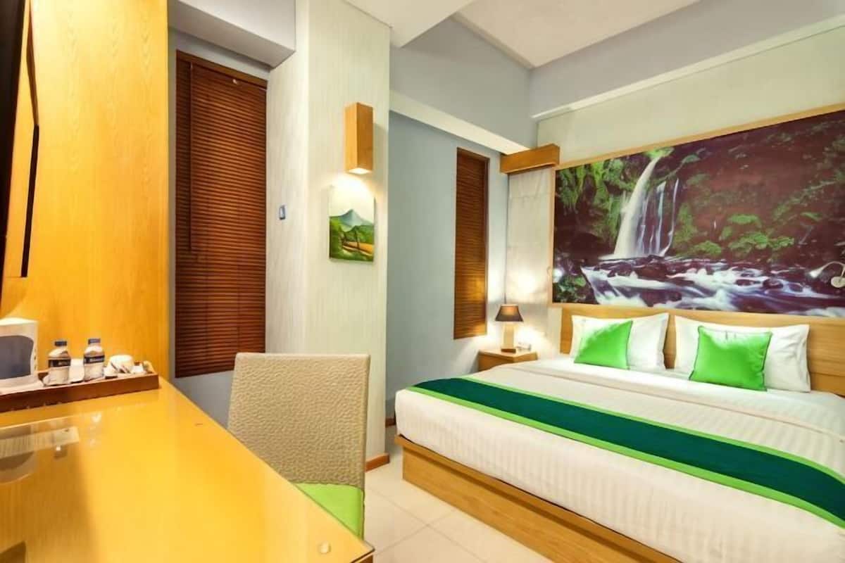 A hotel room with a bed, a desk with a chair, a nightstand, and a painting of a waterfall on the wall.