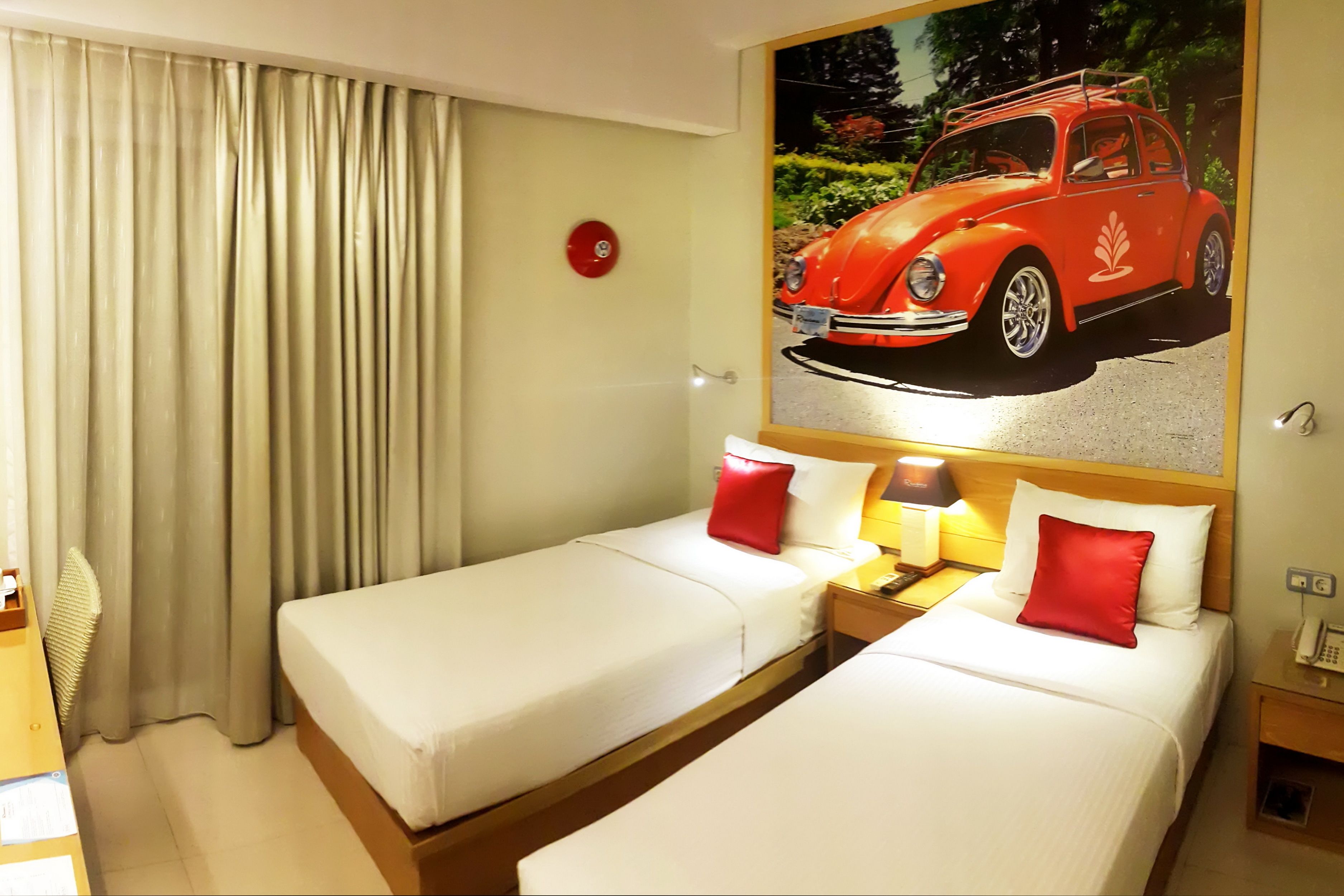 A hotel room with two beds, a red pillow, a nightstand with a lamp, and a wall-mounted picture of a car.