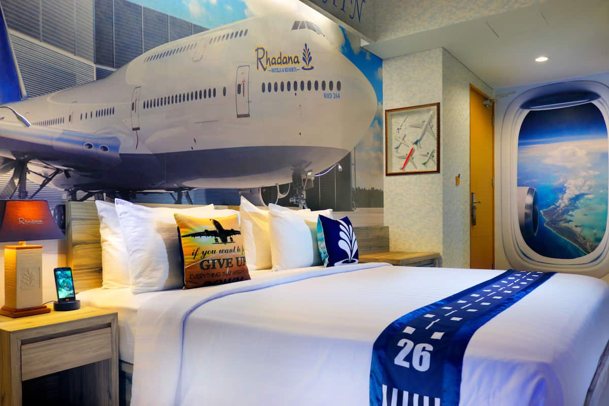 A hotel room with a large airplane model, a bed with a blue and white bedspread, and a window with a view of the sky and clouds.