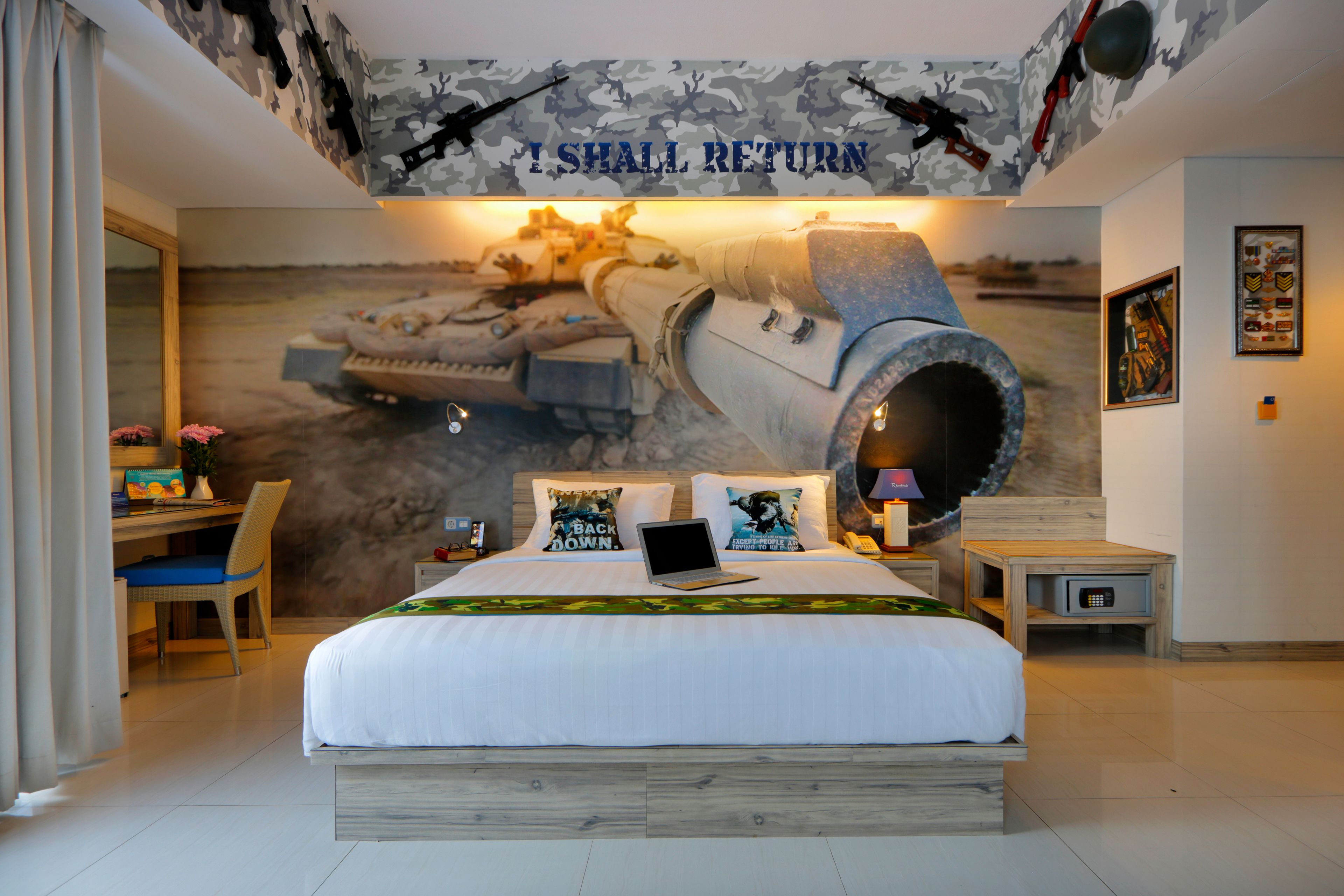 Une chambre avec un grand lit, un bureau et une chaise. Un mur est décoré d’une fresque représentant du matériel militaire.