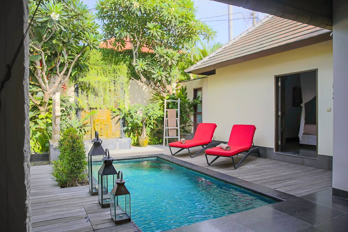 Une piscine bordée de deux chaises longues rouges, entourée de verdure et agrémentée d’un petit jardin avec des lanternes.