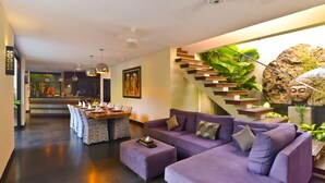 A modern living room with a purple sofa, a dining area with a wooden table and chairs, and a staircase with plants. - Elok Villas (Seminyak)