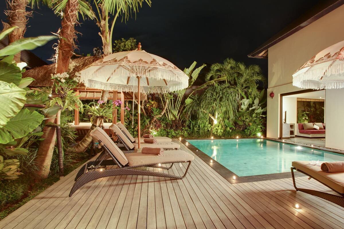 Une terrasse en bois avec des chaises longues et des parasols, une piscine et une végétation tropicale luxuriante.