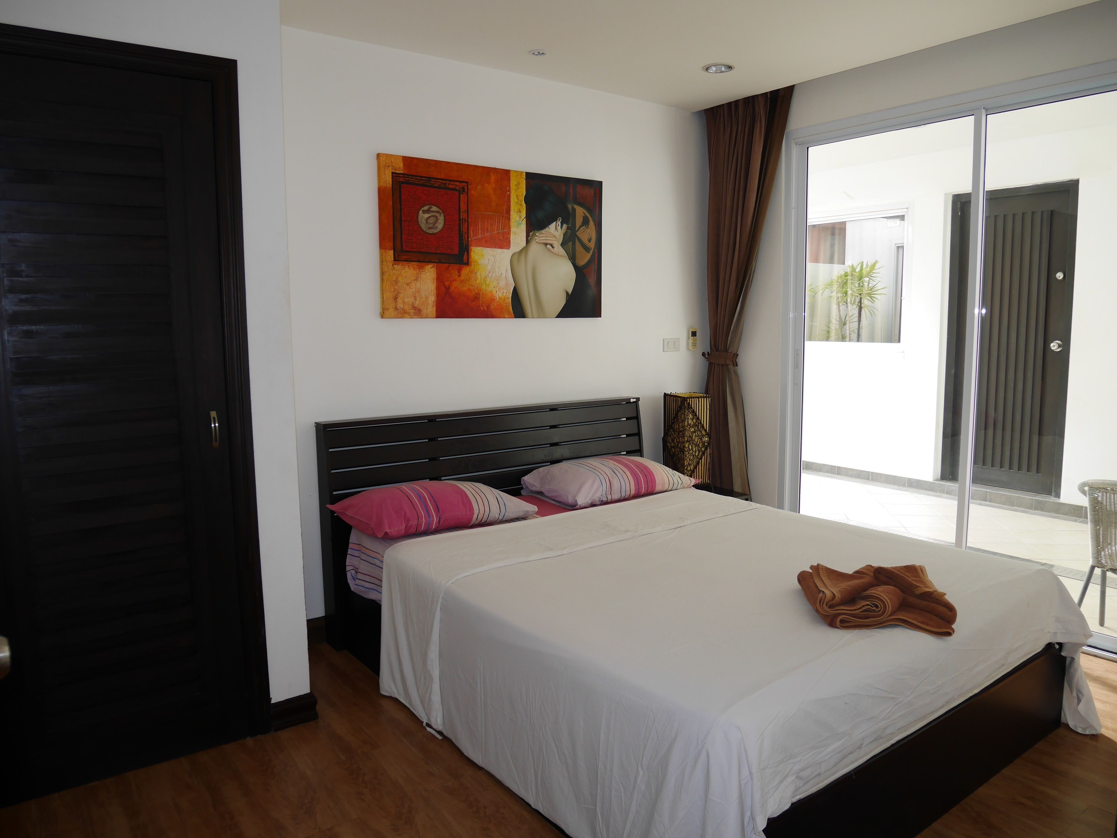 A bedroom with a large bed, a painting on the wall, and a sliding glass door to the outside.