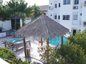 A poolside area with a thatched umbrella, lounge chairs, and a white building in the background. - Tropical Paradise Residence (Koh Samui)