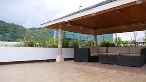 Ein Außenbereich mit Holzterrasse, einem überdachten Bereich mit Dach und Blick auf ein Gebäude sowie Grünflächen im Hintergrund.
