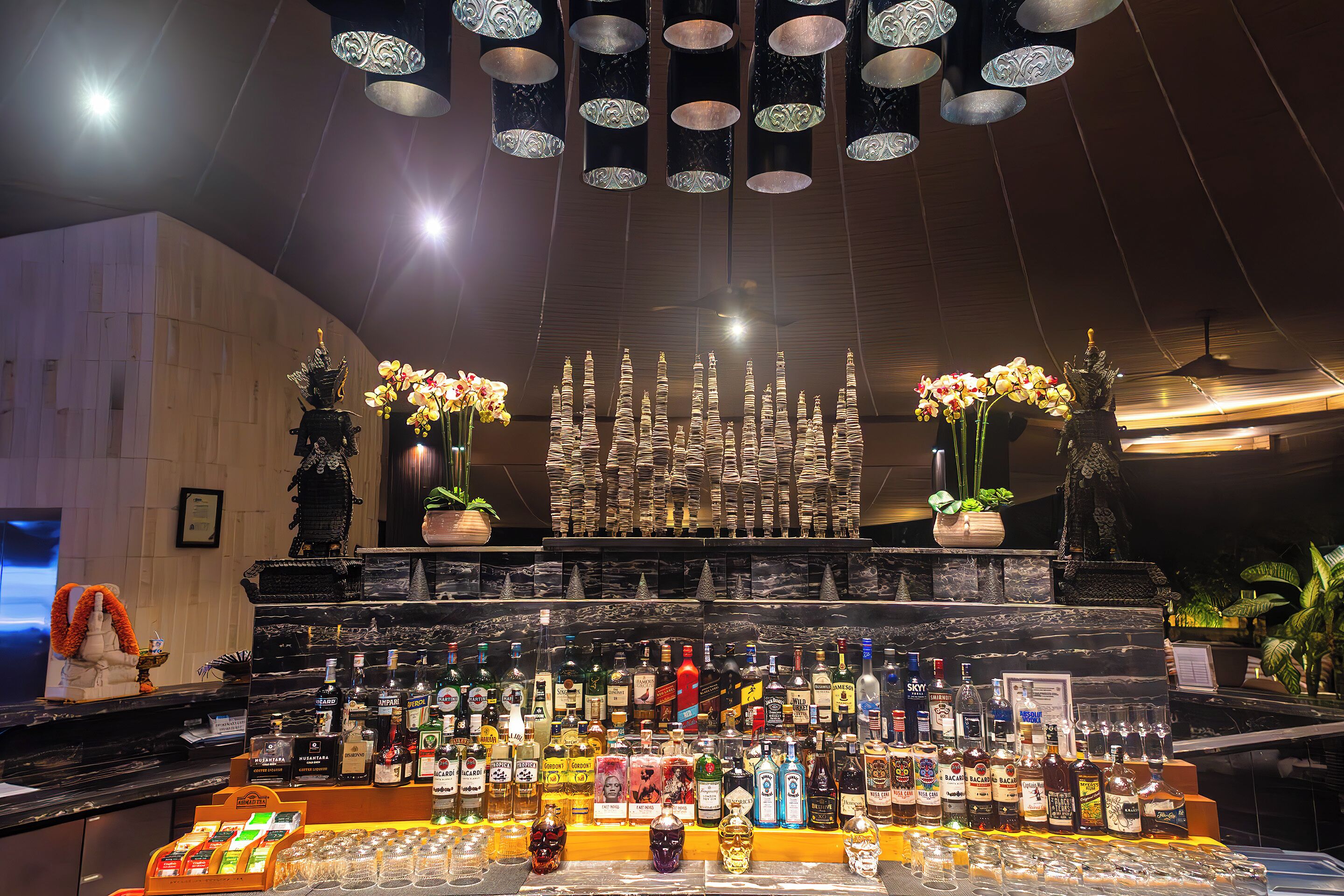 A bar area with a variety of liquor bottles and glasses, decorative vases with flowers, and a patterned ceiling.