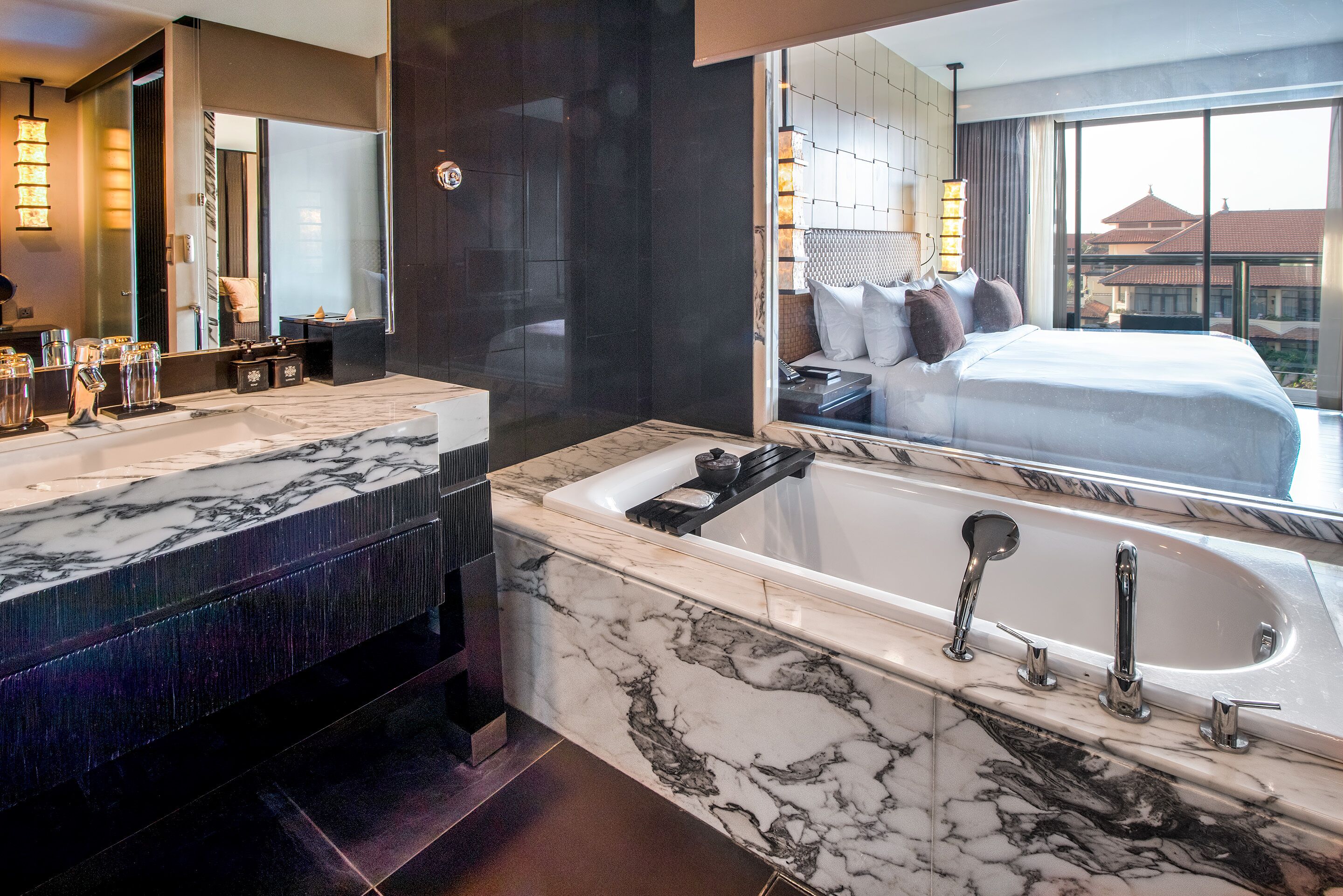 Un baño moderno con encimeras de mármol, una bañera grande y un espejo amplio que refleja un dormitorio con cama y almohadas.