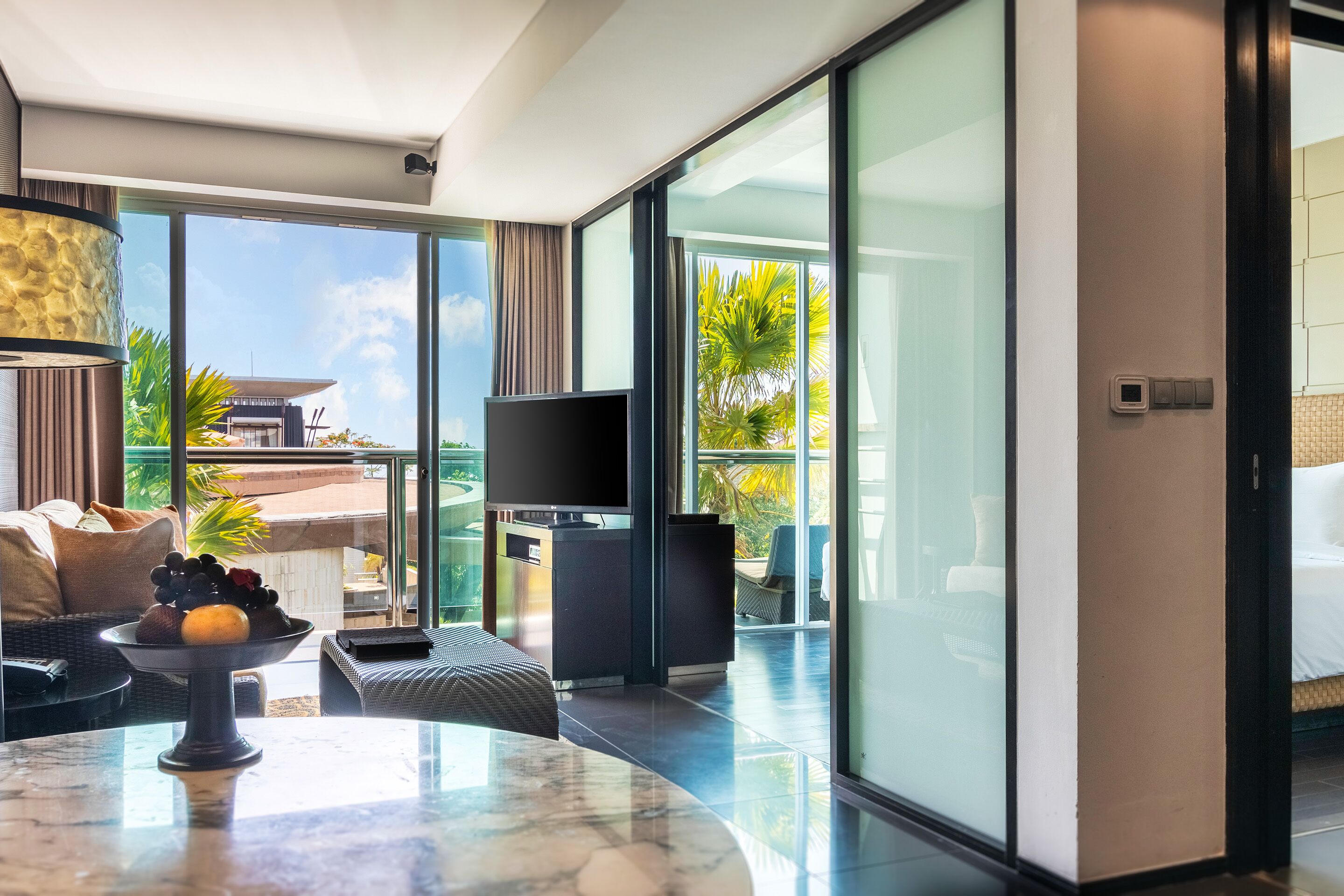 Une chambre d’hôtel moderne dotée d’un grand balcon, d’un téléviseur à écran plat et d’une table en marbre sur laquelle sont disposés des fruits.