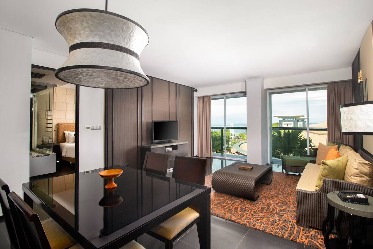 Ein modernes Hotelzimmer mit Essbereich, einer Couch mit Kissen, einem Fernseher und einem großen Fenster mit Blick ins Freie.