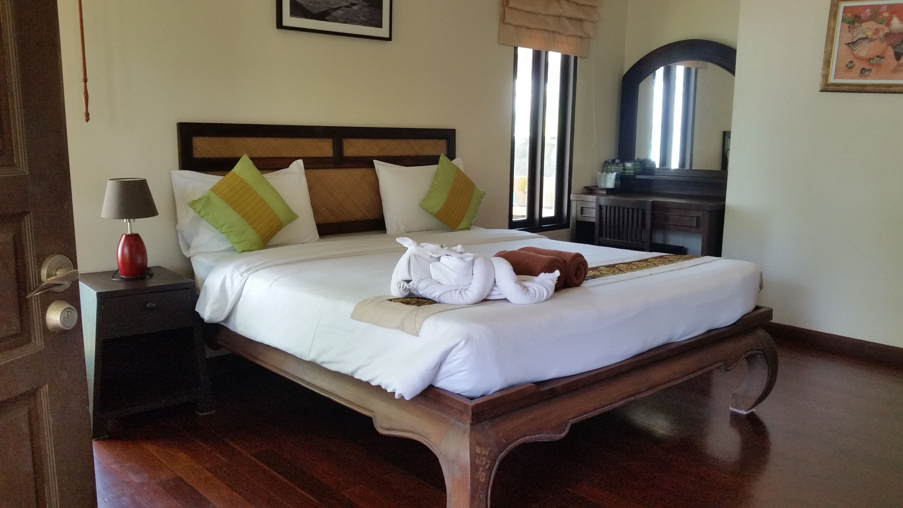 A neatly made bed with white linens, green and yellow pillows, and a brown towel. There is a wooden nightstand with a lamp and a small drawer. A mirror hangs on the wall above the bed.