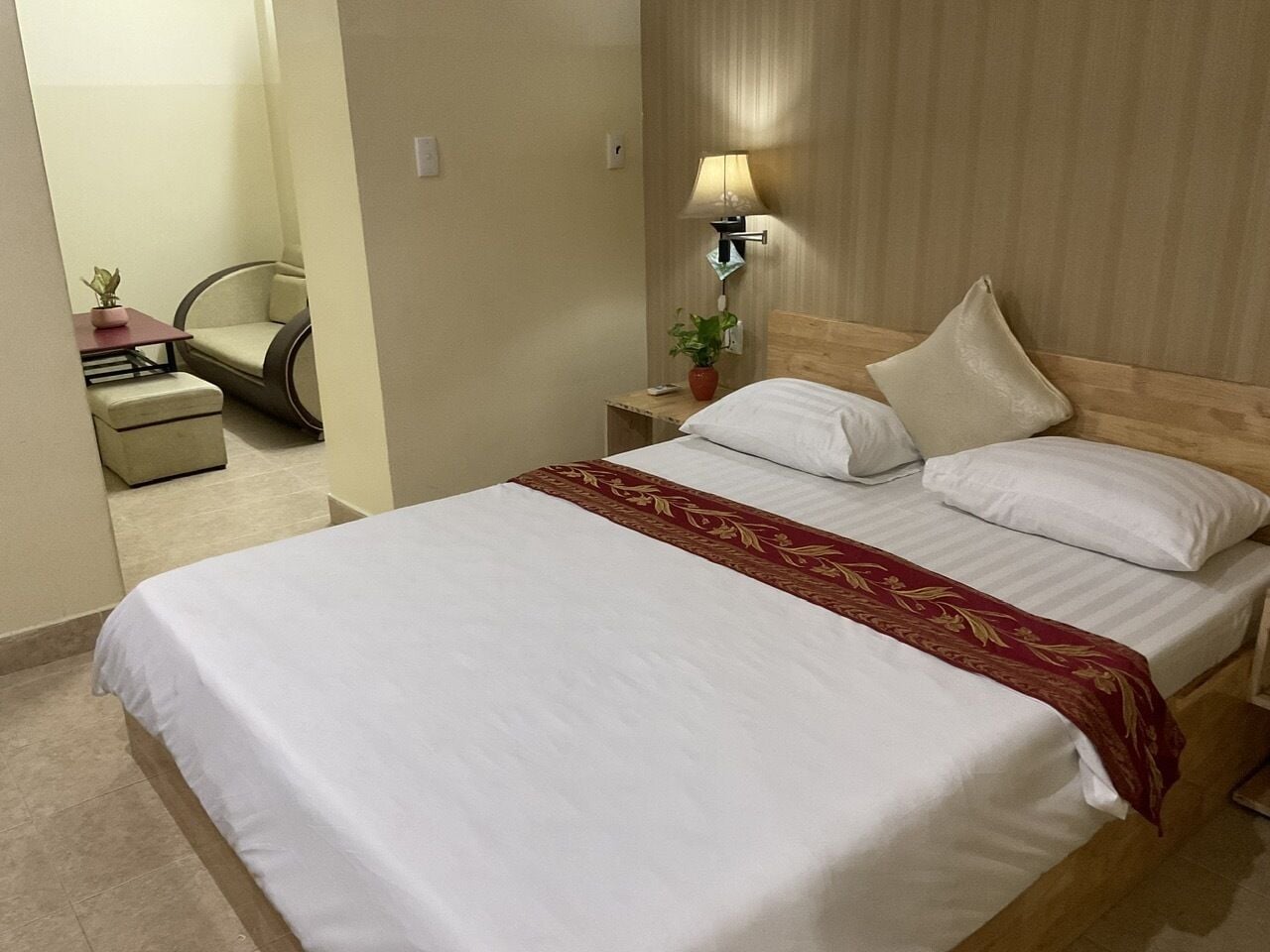A hotel room with a large bed, a bedside table with a lamp, a chair, and a small table with a plant.