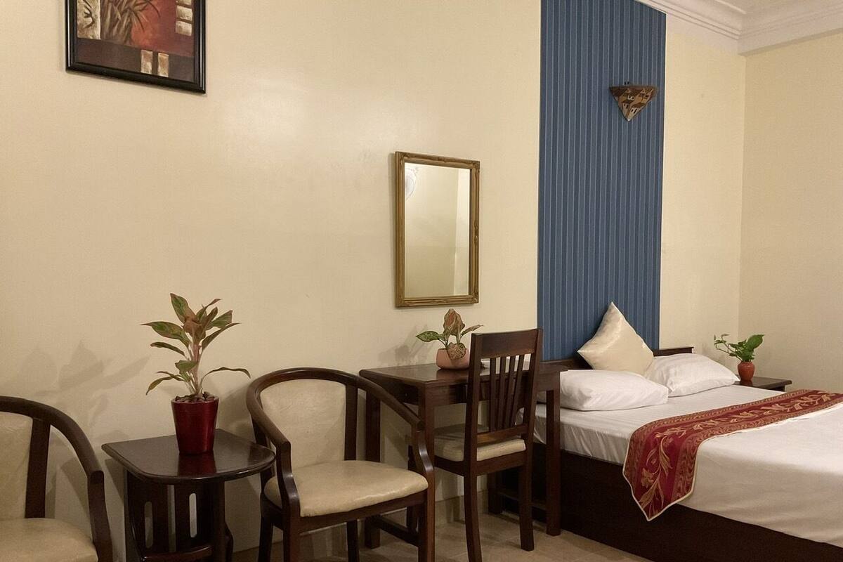 Une chambre d’hôtel avec un lit, deux chaises, une petite table, un miroir et une plante en pot.