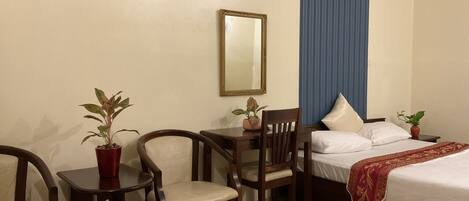 A hotel room with a bed, two chairs, a small table, a mirror, and a potted plant.