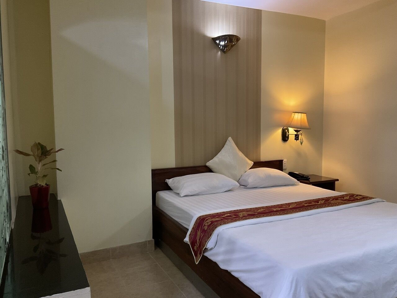 A neatly made bed with white linens and a red decorative throw, a bedside lamp, and a potted plant on a dark wooden nightstand.