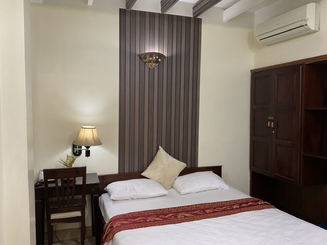 A hotel room with a bed, a desk with a lamp, a wardrobe, and a wall-mounted air conditioner.