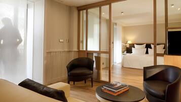 Ein Hotelzimmer mit einer Sitzecke, zwei Sesseln, einem kleinen Tisch mit einem Buch und einem Bett mit weißer Bettwäsche.