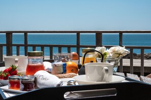 A breakfast setup with a view of the ocean, including a cup, a jar of jam, a bottle of orange juice, and a flower vase.