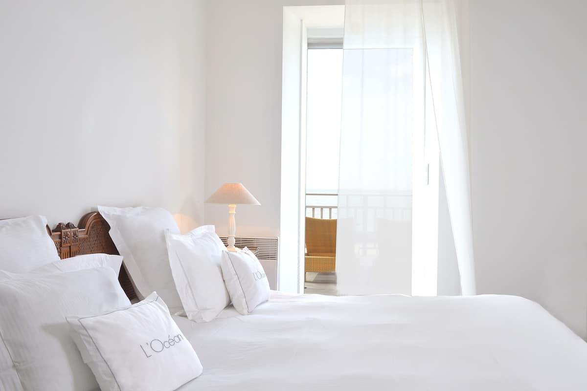 Una cama bien hecha con sábanas y almohadas blancas, una lámpara de noche y vistas a un balcón a través de una ventana con cortinas translúcidas.