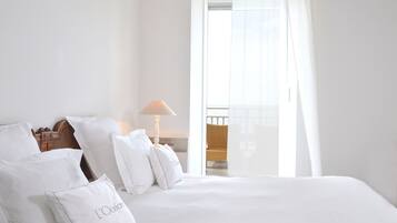 A neatly made bed with white linens and pillows, a bedside lamp, and a view of a balcony through a window with sheer curtains.
