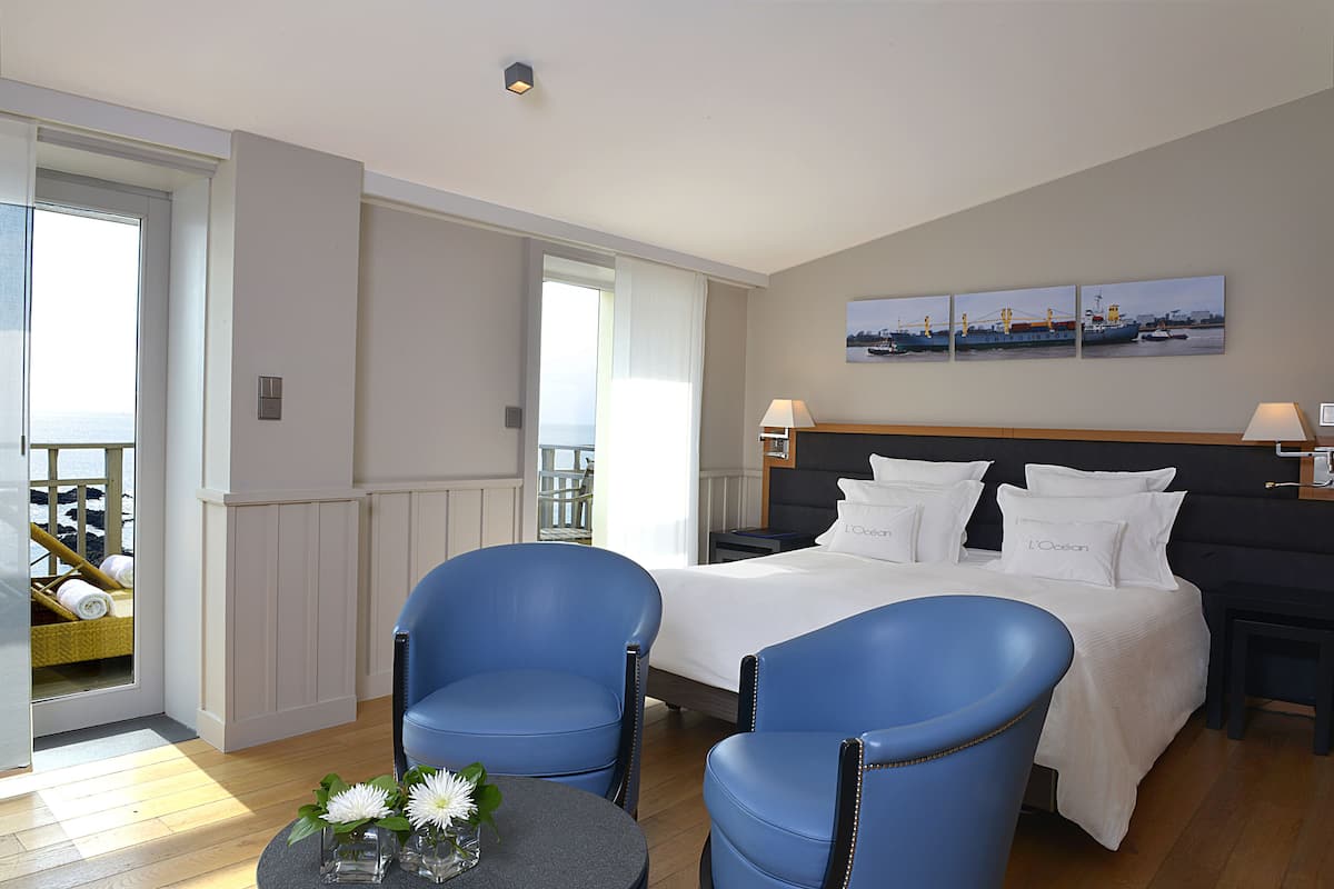 Habitación de hotel con cama, dos sillas azules, una mesita con flores, un balcón con vistas y un cuadro en la pared.