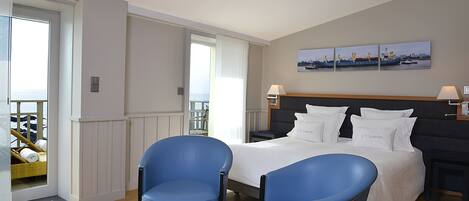 Habitación de hotel con cama, dos sillas azules, una mesita con flores, un balcón con vistas y un cuadro en la pared.