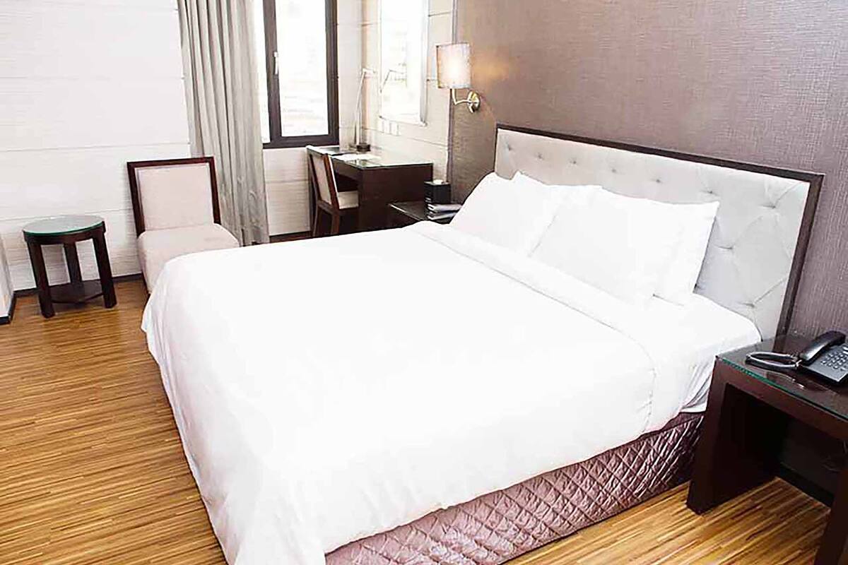 Una cama bien hecha con sábanas blancas, una mesita de noche con lámpara y una silla en una habitación de hotel con suelo de madera y una ventana con cortinas.