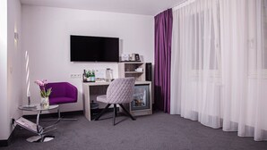 Una habitación de hotel moderna con una televisión de pantalla plana, un sillón morado, una mesa de centro de vidrio, una silla blanca, un minibar y cortinas de voile blancas.