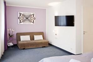 Un dormitorio con un sofá marrón, un televisor de pantalla plana montado en la pared, una planta en maceta y una obra de arte enmarcada.