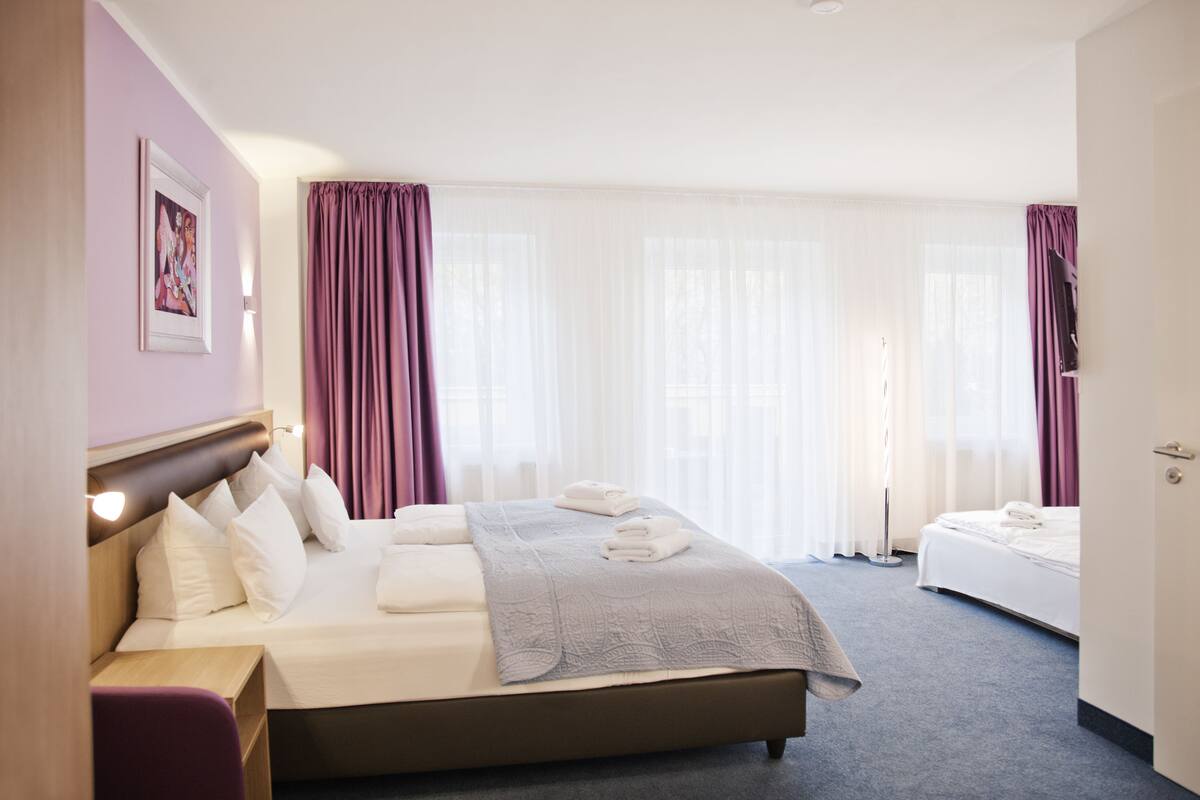 Ein Hotelzimmer mit einem großen Bett, einem Nachttisch, einem Sessel und einem Fenster mit Vorhängen.