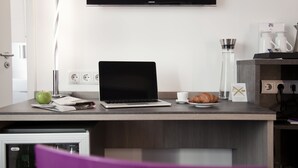 Una habitación de hotel moderna con un escritorio de madera, una computadora portátil, un croissant, una manzana verde, una taza y un televisor de pantalla plana montado en la pared.