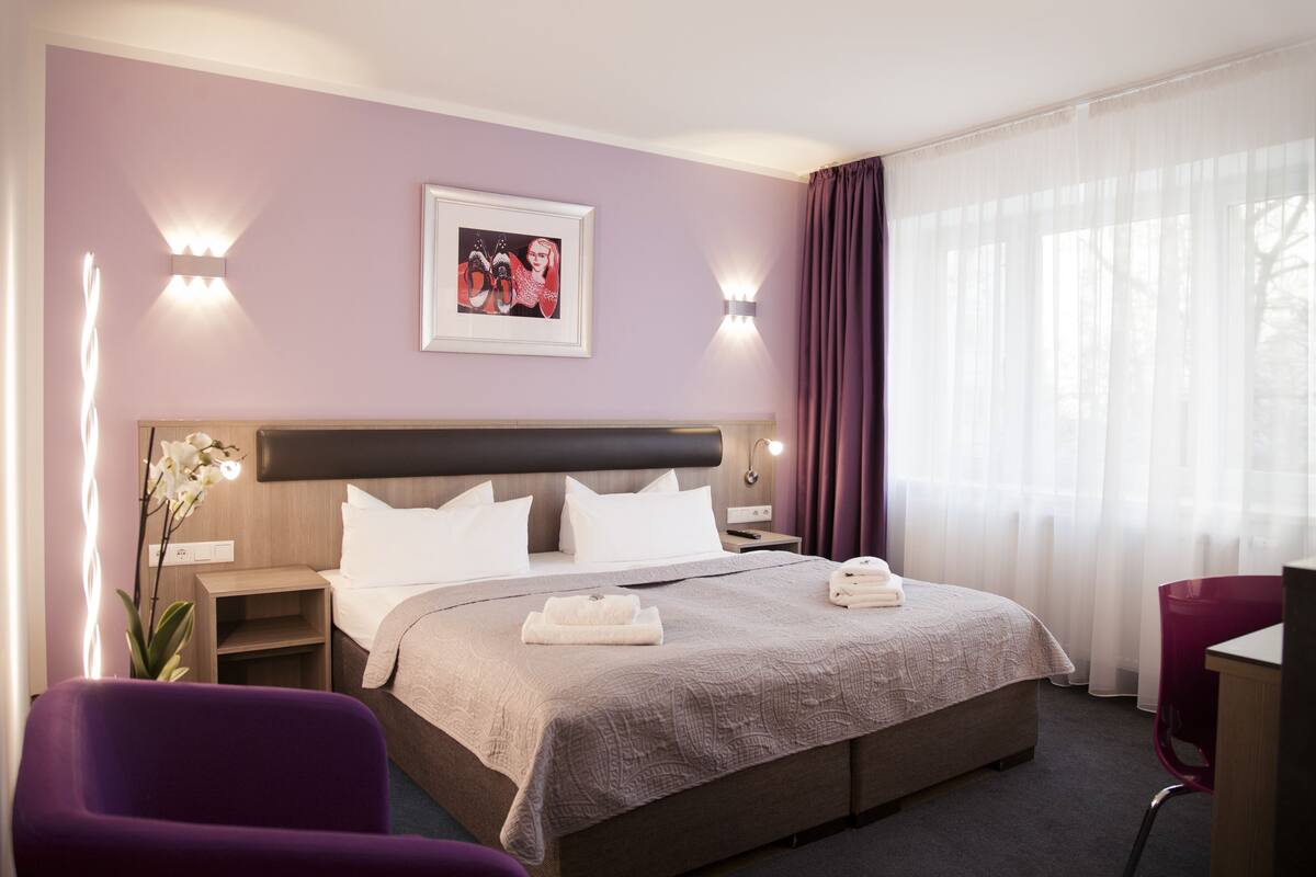 Ein Hotelzimmer mit einem großen Bett, einem Nachttisch, einem Schreibtisch, einem Stuhl und einem Fenster mit Vorhängen.