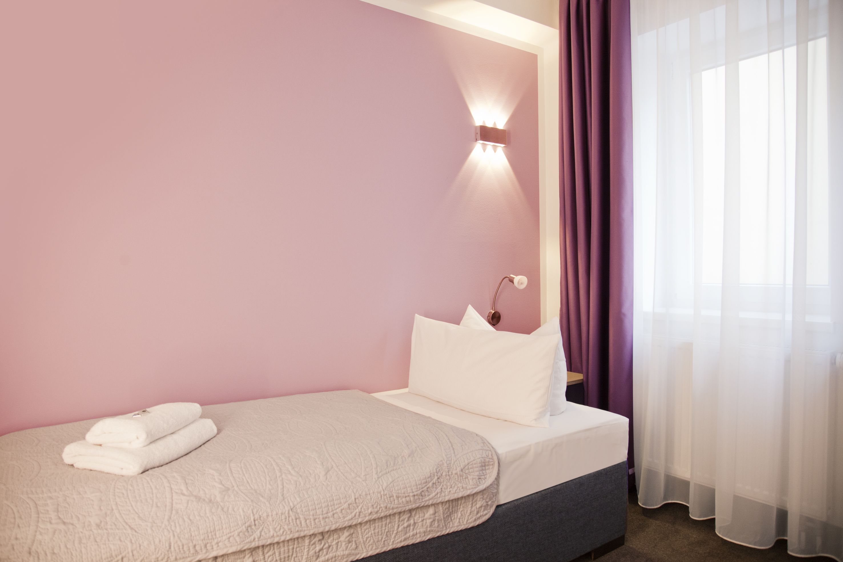 Quarto com uma cama de solteiro, luminária de cabeceira e janela com cortinas finas.
