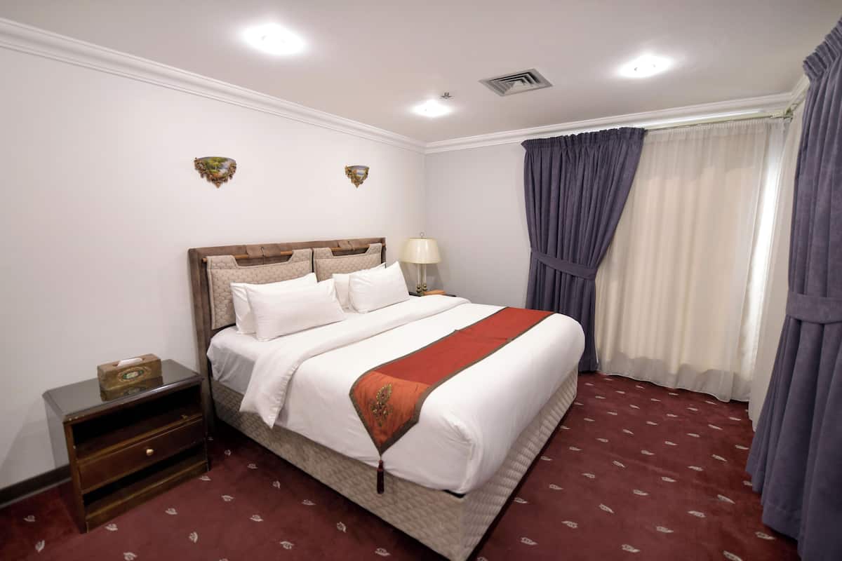 A neatly made bed with white linens and a brown headboard, a bedside table with a lamp, and a patterned carpet.