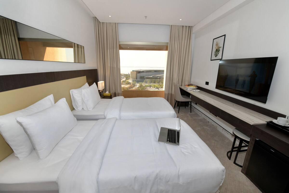 A hotel room with two beds, a desk, a television, and a view of the city.