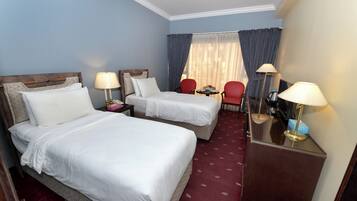 Habitación de hotel con dos camas, un escritorio, dos sillas, una ventana con cortinas y una alfombra roja.