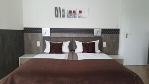 Una cama doble con una colcha marrón, dos almohadas blancas y un cabecero gris. Hay dos mesitas de noche con lámparas blancas. La pared tiene un diseño de baldosas blanco y negro.