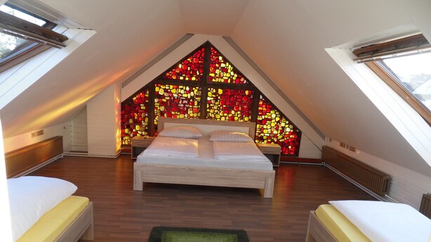 Quarto com teto inclinado, cama, mesa de cabeceira e parede com vitrais coloridos.